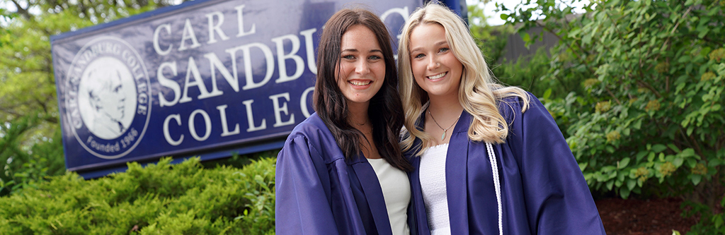 two female students in graduation gowns standing in front of the college sign.