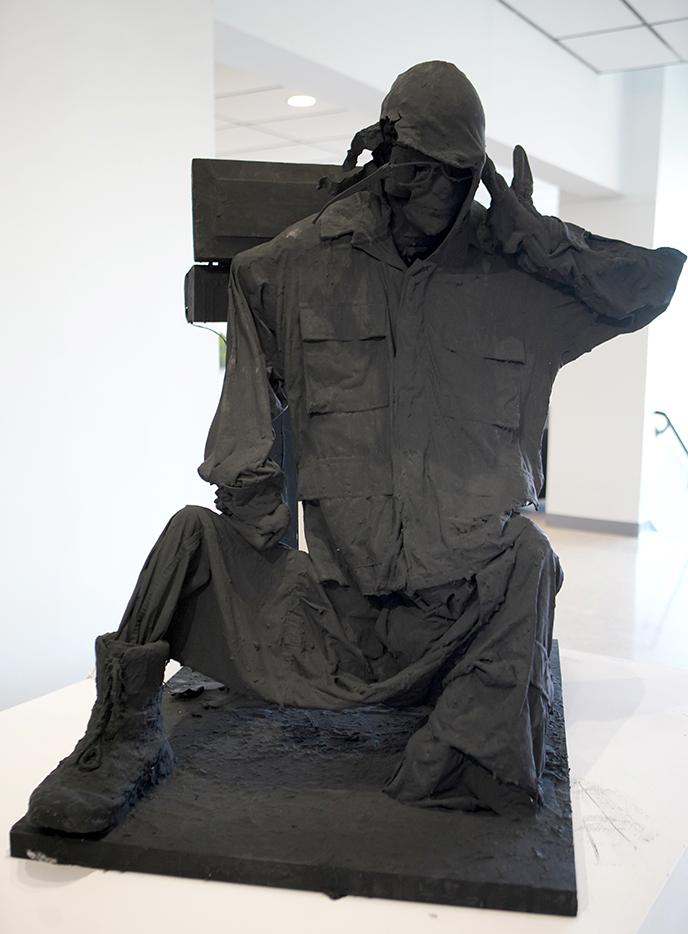 Sculpture, a figure dressed in military clothing painted black saluting. sitting on the ground