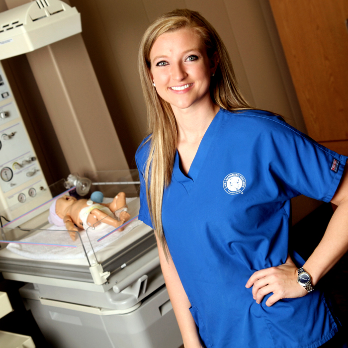 a women standing in front of a medical equipment holding a simulation baby. The women is wearing blue scrubs.