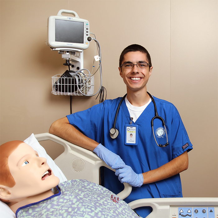 male nursing student standing next to a hospital bed. In the bed is a simulation patient.