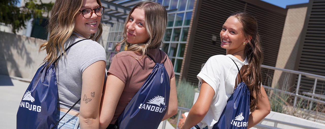 Three female students looking over their sholder at the camera with Sandburg backpacks on.