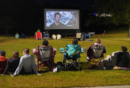 people sitting in grass watching a large screen with a movie on it.